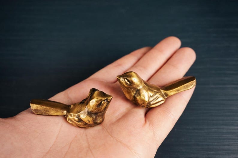 May include: Two small, gold-colored bird figurines. The birds are detailed with visible wings and beaks. They are resting on a person's open palm, with a dark blue background. The birds appear to be made of metal.
