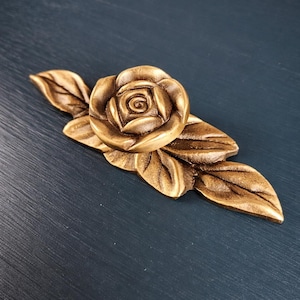 May include: A bronze-colored decorative rose and leaf embellishment. The rose is in full bloom, with detailed petals and a central spiral. The leaves are elongated and detailed. The piece is likely for furniture or craft projects.
