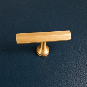 May include: A gold-toned cabinet pull with a hexagonal bar and a round base. The hardware is set against a dark blue background. This decorative pull is suitable for drawers and cabinets.