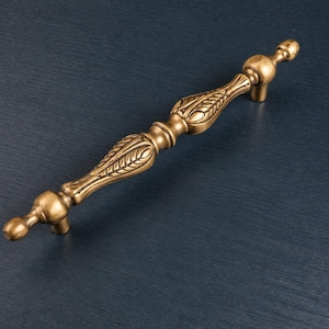 May include: A gold-toned cabinet pull with an ornate design. The handle features a central section with leaf-like details and rounded ends. The hardware is set against a dark blue background.