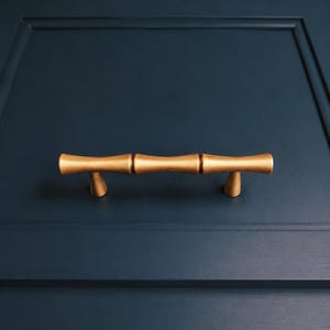 May include: Gold-colored bamboo-style cabinet pulls mounted on a navy blue cabinet door. The pulls have a textured surface and a cylindrical shape, adding a touch of natural elegance to the furniture. The hardware is centered on the door.
