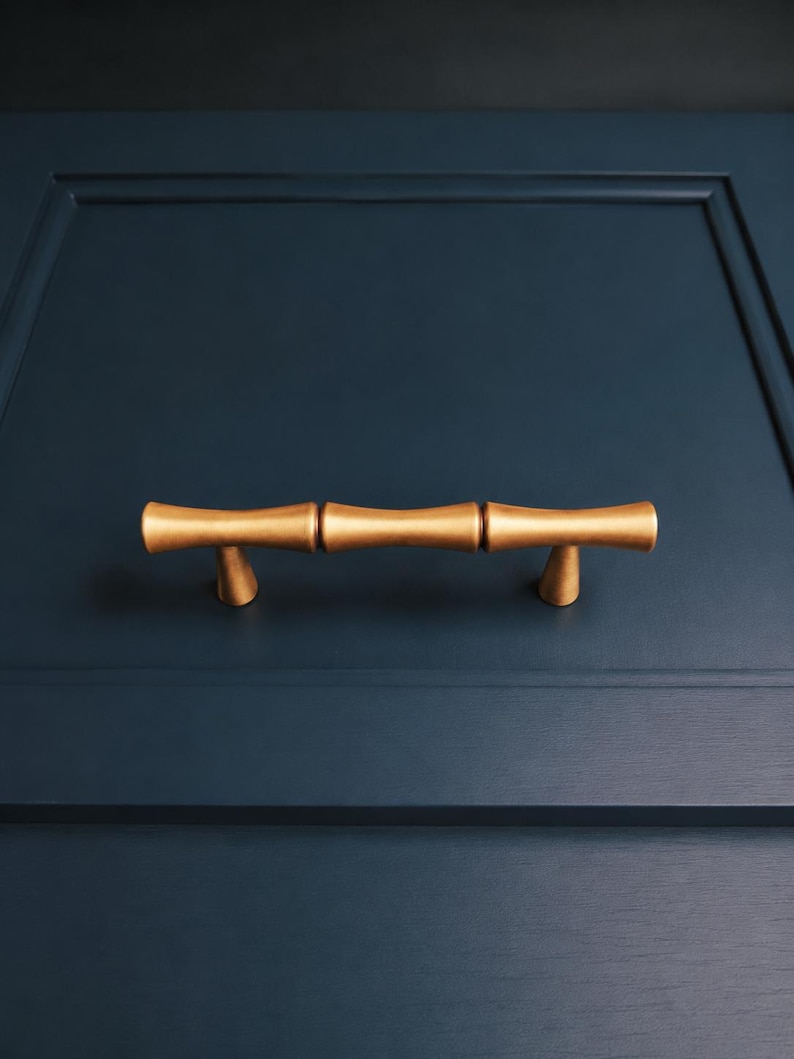 May include: Gold-colored bamboo-style cabinet pulls mounted on a navy blue cabinet door. The pulls have a textured surface and a cylindrical shape, adding a touch of natural elegance to the furniture. The hardware is centered on the door.