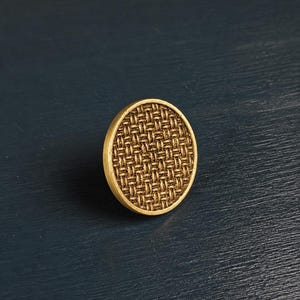 May include: A circular brass cabinet knob with a woven pattern. The knob has a raised, circular design and a warm, metallic finish. Suitable for furniture and home decor projects. The diameter is approximately 4 cm.