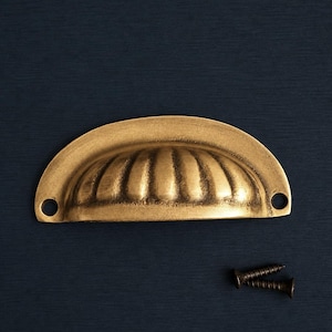 Könnte beinhalten: Ein goldfarbener, halbmondförmiger Schubladengriff mit geripptem Design. Zwei dunkle Schrauben sind darunter positioniert. Die Hardware befindet sich vor einem dunkelblauen Hintergrund.