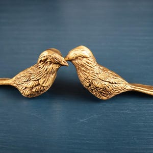 May include: Two gold-colored bird figurines, facing each other, with their beaks touching. The birds have detailed feather textures and are set against a dark blue background. Decorative home decor.