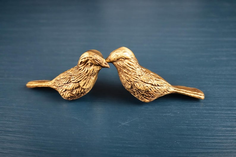 May include: Two gold-colored bird figurines, facing each other, with their beaks touching. The birds have detailed feather textures and are set against a dark blue background. Decorative home decor.
