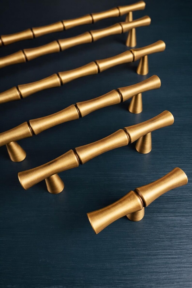May include: Gold-colored bamboo-style cabinet pulls arranged on a dark blue surface. The pulls have a textured, segmented design, resembling bamboo stalks. Each pull is mounted on a small, triangular base, providing a decorative and functional hardware option.