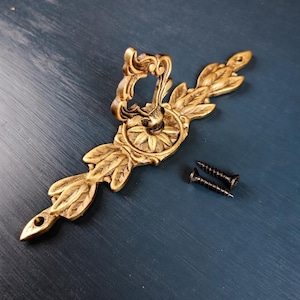 May include: Ornate brass-toned furniture handle with leaf and floral detailing. Includes two black screws. The handle has a decorative design, perfect for adding a vintage touch to cabinets or drawers.