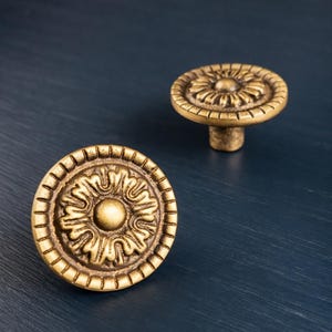 Puede incluir: Dos tiradores de armario de latón antiguo con diseño floral. Los tiradores son redondos con un patrón detallado y un pequeño vástago. El fondo es de color azul oscuro.
