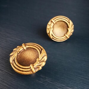 May include: Two antique-style, gold-colored cabinet knobs. Each knob features a circular design with a central recessed area and an outer ring adorned with leaf-like details. The knobs are set against a dark blue background.