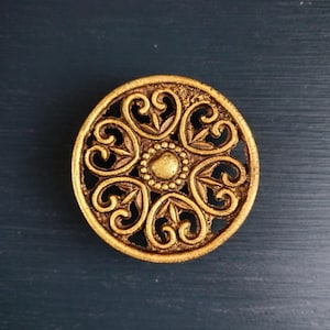 Op de afbeelding: Een rond, antiek ogend, goudkleurig decoratief object. Het object heeft een ingewikkeld openwerkontwerp met hart- en bladvormen die uitstralen van een centraal, verhoogd, cirkelvormig element. De algehele esthetiek is sierlijk en vintage.
