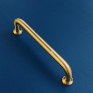 May include: A gold-toned cabinet pull handle with a curved bar and rounded ends. The handle is set against a deep blue background, creating a contrast that highlights its design. This hardware is suitable for drawers and cabinets.