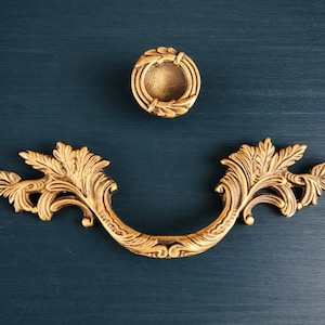 Puede incluir: Juego de herrajes decorativos dorados, que incluye un pomo redondo y un tirador curvo. Los herrajes presentan diseños ornamentados de hojas y flores, sobre un fondo azul verdoso oscuro. Ideal para la restauración de muebles.