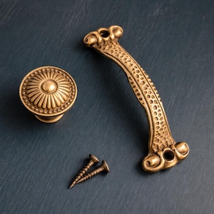 May include: Antique brass-colored cabinet hardware set, including a round knob with a sunburst design, a pull handle with a textured pattern, and two screws. The hardware is displayed on a dark blue surface.