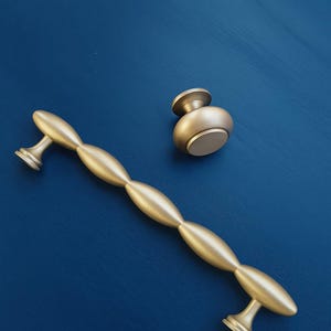 Pode incluir: Conjunto de ferragens douradas para armários, incluindo uma puxador longo e um puxador redondo. O puxador apresenta um design alongado e com contas, enquanto o puxador tem formato de cogumelo. Ambas as peças estão sobre um fundo azul escuro.