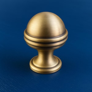May include: A brass-toned cabinet knob with a spherical top and a decorative, ridged band. The knob sits on a pedestal base, showcasing a classic design. The hardware is set against a deep blue background.