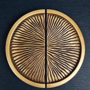 Puede incluir: Dos mitades de un tirador de armario circular dorado con un diseño texturizado de rayos de sol. Los tiradores se colocan sobre un fondo azul oscuro, mostrando los intrincados detalles y el acabado metálico.