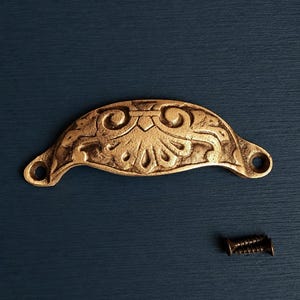 Könnte beinhalten: Ein goldfarbener, verzierter Schubladengriff mit geschwungenem Design und dekorativen floralen Mustern. Inklusive passender Schraube zur Montage. Die Hardware befindet sich vor einem dunkelblauen Hintergrund.