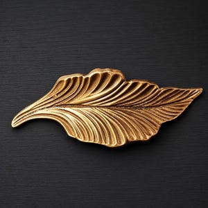 May include: A gold-colored, leaf-shaped decorative object with detailed veining. The object has a textured surface and is set against a dark background. The leaf design is a common motif in home decor and art.
