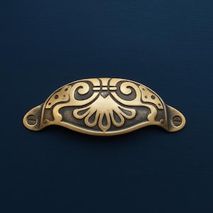 May include: An antique-style brass drawer pull with an intricate floral pattern. The pull is curved and features two screw holes. The brass has a slightly aged look. The background is a dark blue. The pull is approximately 12 cm in length.