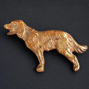 Peut inclure: Une figurine de chien de couleur dorée en position debout, la gueule ouverte. Le chien a une texture de fourrure détaillée et est posé sur un fond sombre. La sculpture semble être en métal.
