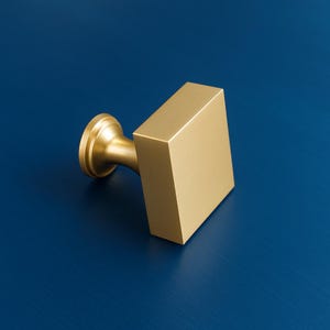May include: A gold-coloured cabinet knob with a square handle and a circular base. The knob is set against a deep blue background, highlighting its metallic finish and geometric design. This hardware piece adds a touch of modern elegance.