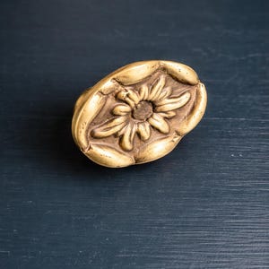 May include: A golden-colored, oval-shaped decorative knob with a raised floral design. The knob features a detailed sunflower in the center, surrounded by a textured border. The background is a dark blue surface.