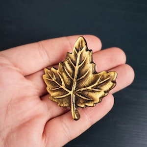 May include: A detailed, golden-colored leaf-shaped decorative object. The leaf has intricate vein detailing and a slightly textured surface. The object is likely made of metal and has a vintage or antique appearance. It is a decorative element.