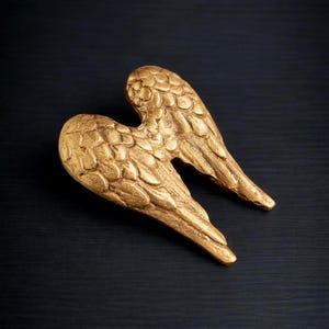 May include: A pair of golden angel wings, with detailed feather textures, are displayed against a dark background. The wings are positioned to appear as if they are in flight, creating a sense of movement and grace. The wings are a decorative item.