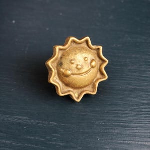 Pode incluir: Um puxador de armário em forma de sol, cor de latão, com um rosto sorridente. O puxador tem uma superfície texturizada e detalhada e um design de estrela ao redor do rosto. O hardware é provavelmente para uma gaveta ou armário.