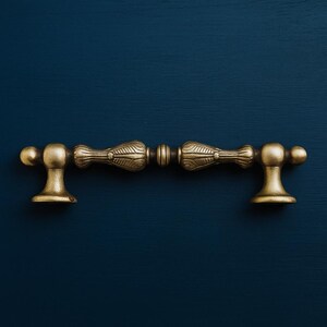 May include: Two antique brass cabinet pulls. Each pull has a decorative central section with an etched design, with rounded ends and a small, circular base. The hardware is set against a dark blue background.