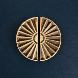 Puede incluir: Dos mitades de un tirador de armario de latón, cada una con forma semicircular. Los tiradores presentan un diseño de sol con rayos triangulares que irradian de un círculo central. El fondo es azul oscuro.