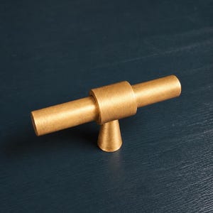 May include: A gold-colored cabinet pull with a cylindrical bar and a conical base. The hardware is made of metal and is set against a dark blue background. This modern style pull is suitable for drawers and cabinets.