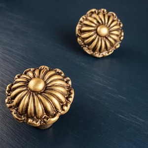 May include: Two antique-style brass cabinet knobs with a floral design. Each knob features a central, raised circle surrounded by radiating lines and a decorative edge. The knobs are set against a dark blue background.