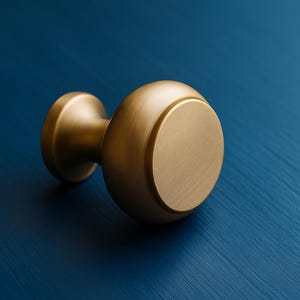 Puede incluir: Un pomo de armario dorado con un diseño redondeado. El pomo tiene una cara circular y una base cilíndrica, sobre un fondo azul. El herraje es probablemente para muebles o armarios.
