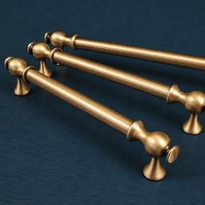May include: Four antique brass cabinet pulls with a classic design. Each pull features a rounded end and a decorative base, perfect for adding a vintage touch to furniture. The pulls are arranged diagonally against a dark blue background.