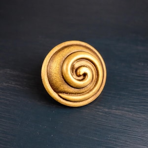 May include: A golden-colored, circular cabinet knob with a spiral design. The knob has a slightly textured surface and is set against a dark blue background. This decorative hardware adds a touch of elegance to furniture.