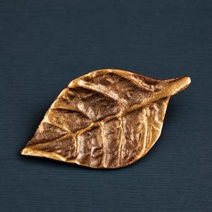 Op de afbeelding: Een bronzen, bladvormig decoratief object. Het blad heeft gedetailleerde nerven en een getextureerd oppervlak, met een lichte kromming. Het object staat tegen een donkerblauwe achtergrond, waardoor een contrast ontstaat dat de vorm van het blad benadrukt.