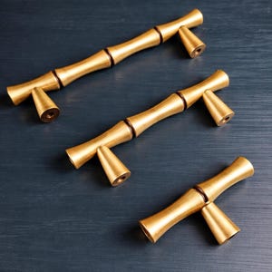 May include: Three gold-colored bamboo-style cabinet pulls with a dark brown accent at the joints. The pulls have a cylindrical body and conical ends. The hardware is arranged on a dark blue surface.