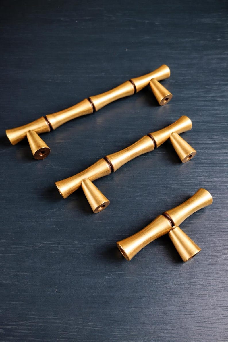 May include: Three gold-colored bamboo-style cabinet pulls with a dark brown accent at the joints. The pulls have a cylindrical body and conical ends. The hardware is arranged on a dark blue surface.
