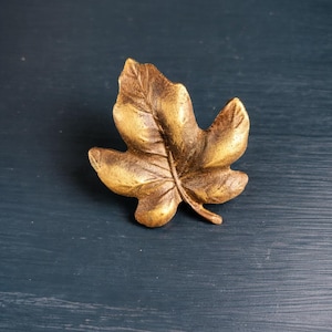 Peut inclure: Un objet décoratif en forme de feuille de couleur bronze. La feuille présente des nervures détaillées et une surface texturée. L'objet est probablement une poignée de meuble ou un élément décoratif, et il est posé sur un fond bleu foncé.
