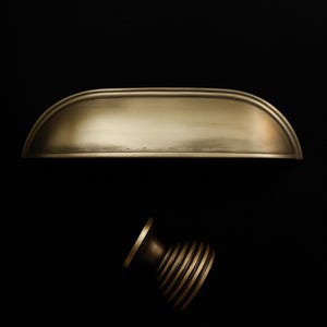 May include: Two brass-toned cabinet hardware pieces against a black background. One is a long, curved pull, and the other is a round knob with horizontal grooves. The hardware has a vintage aesthetic.