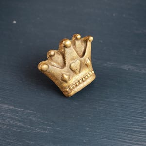 Pode incluir: Uma pequena coroa dourada com um detalhe em forma de coração. A coroa é feita de um material texturizado e tem uma aparência ligeiramente envelhecida. A coroa é um item decorativo, possivelmente para um topo de bolo ou joia.