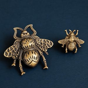 May include: Two brass-colored insect-shaped decorative objects. One is a large, detailed beetle with outstretched wings. The other is a smaller bee with detailed wings. Both are on a dark blue background.