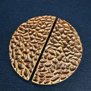 May include: Two halves of a bronze-coloured, textured, circular cabinet pull. The pulls have a hammered, organic design and are set against a dark blue background. The pulls are designed to be installed on a cabinet or drawer.