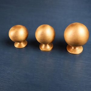 Könnte beinhalten: Drei goldfarbene, kugelförmige Schrankknöpfe mit gebürsteter Oberfläche. Jeder Knopf hat einen abgerundeten Sockel und eine glatte, metallische Oberfläche. Die Knöpfe sind in einer Reihe auf einer dunkelblauen Oberfläche angeordnet.
