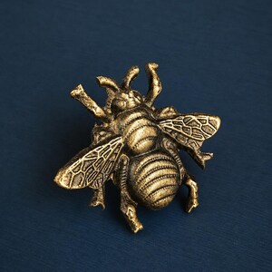 K&ouml;nnte beinhalten: Detaillierte, goldfarbene Bienenfigur mit strukturierten Fl&uuml;geln und K&ouml;rper. Die Biene befindet sich auf einem dunkelblauen Hintergrund. Die Fl&uuml;gel des Insekts sind ausgebreitet, und der K&ouml;rper hat ein gestreiftes Muster.
