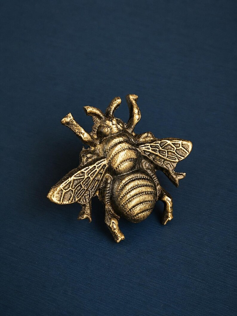 K&ouml;nnte beinhalten: Detaillierte, goldfarbene Bienenfigur mit strukturierten Fl&uuml;geln und K&ouml;rper. Die Biene befindet sich auf einem dunkelblauen Hintergrund. Die Fl&uuml;gel des Insekts sind ausgebreitet, und der K&ouml;rper hat ein gestreiftes Muster.