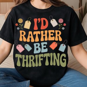 May include: Black t-shirt with the text "I'd Rather Be Thrifting" in a retro font. The design includes colorful tags and flowers. A casual, vintage-inspired tee for those who love thrifting and vintage finds.