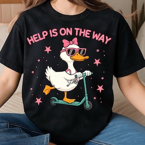 Puede incluir: Camiseta negra con la frase rosa "HELP IS ON THE WAY". El dise&ntilde;o presenta un pato de dibujos animados con gafas de sol y un lazo, montado en un patinete turquesa, con estrellas rosas.
